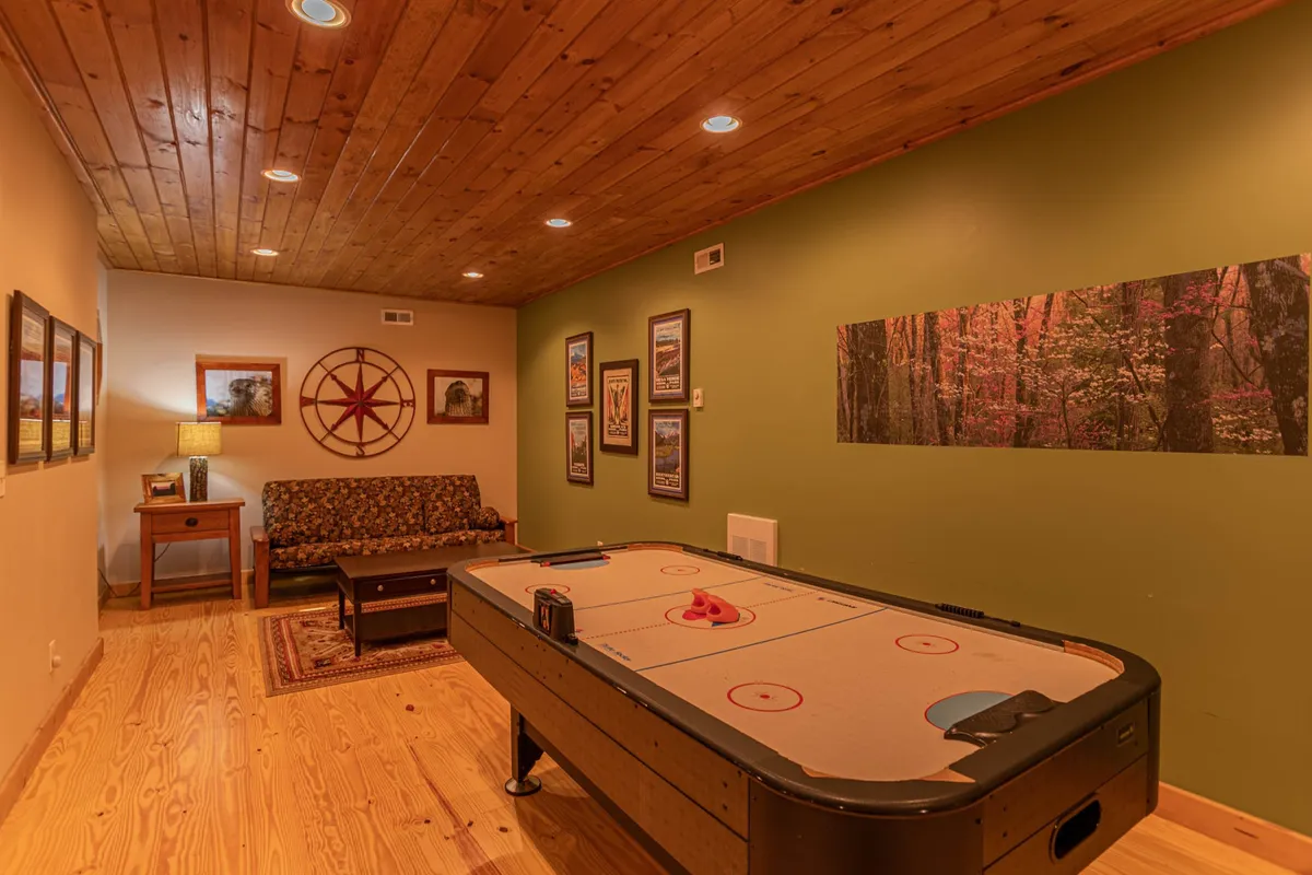 The photo features a vibrant indoor game area with an air hockey table, a comfortable sofa, wall art, and warm lighting from lamps. The space is casual and inviting, perfect for friendly rivalry and family fun in the comfort of the cabin’s cozy interior.