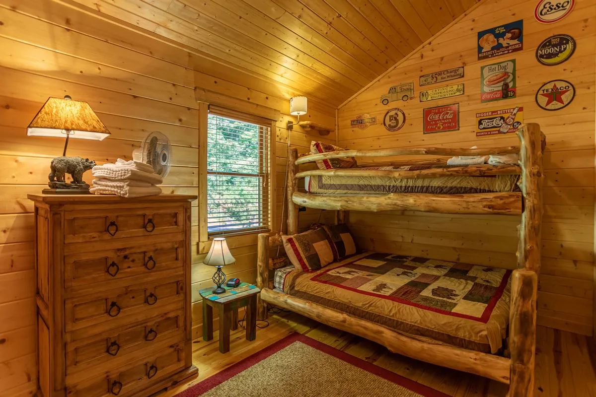 A charming bunk room with Queen-over-Twin bunk beds, a wooden dresser, cozy lighting, and playful vintage signs on the wall. The arrangement makes the space inviting for children or friends sharing a room.