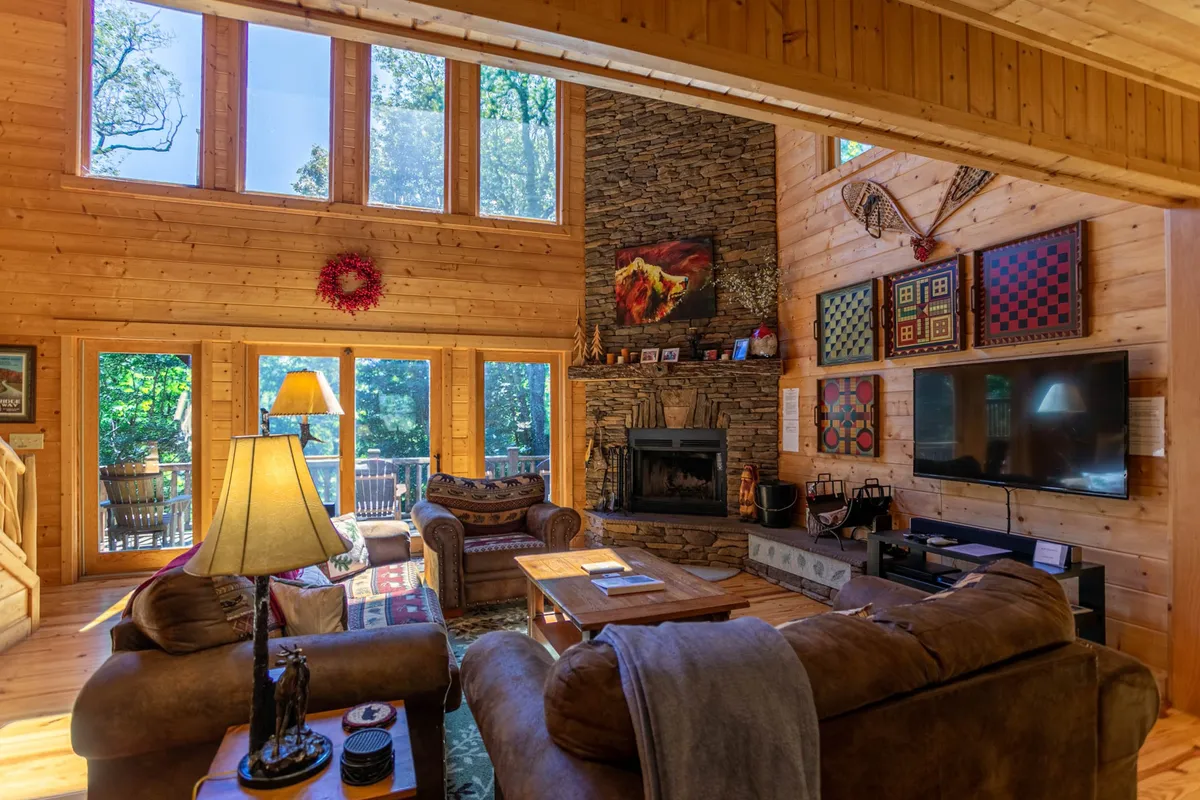 The image showcases the inviting interior of Acorn Lodge, with sunlight streaming through expansive windows, a majestic stone fireplace as the centerpiece, and plush seating arranged for conversation and relaxation. Tasteful wall art and cozy accents create a warm, welcoming atmosphere—perfect for gatherings or quiet evenings in the mountains.