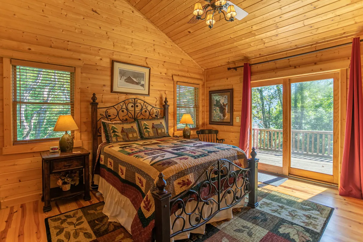 A bright bedroom with a decorative quilt and large windows letting in sunlight, along with a nightstand and lamp. The windows suggest possible views and easy access to the outside, ideal for enjoying morning coffee with mountain views.