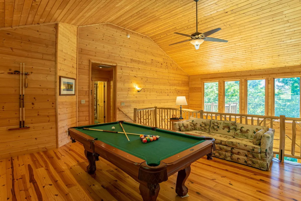 An inviting loft interior featuring a pool table, cozy sofa, large windows with scenic views, and a wooden ceiling. The space is designed for family fun, game nights, and socializing, creating an atmosphere that balances relaxation and excitement. Natural light streams in, underscoring the connection to the beautiful outdoors.