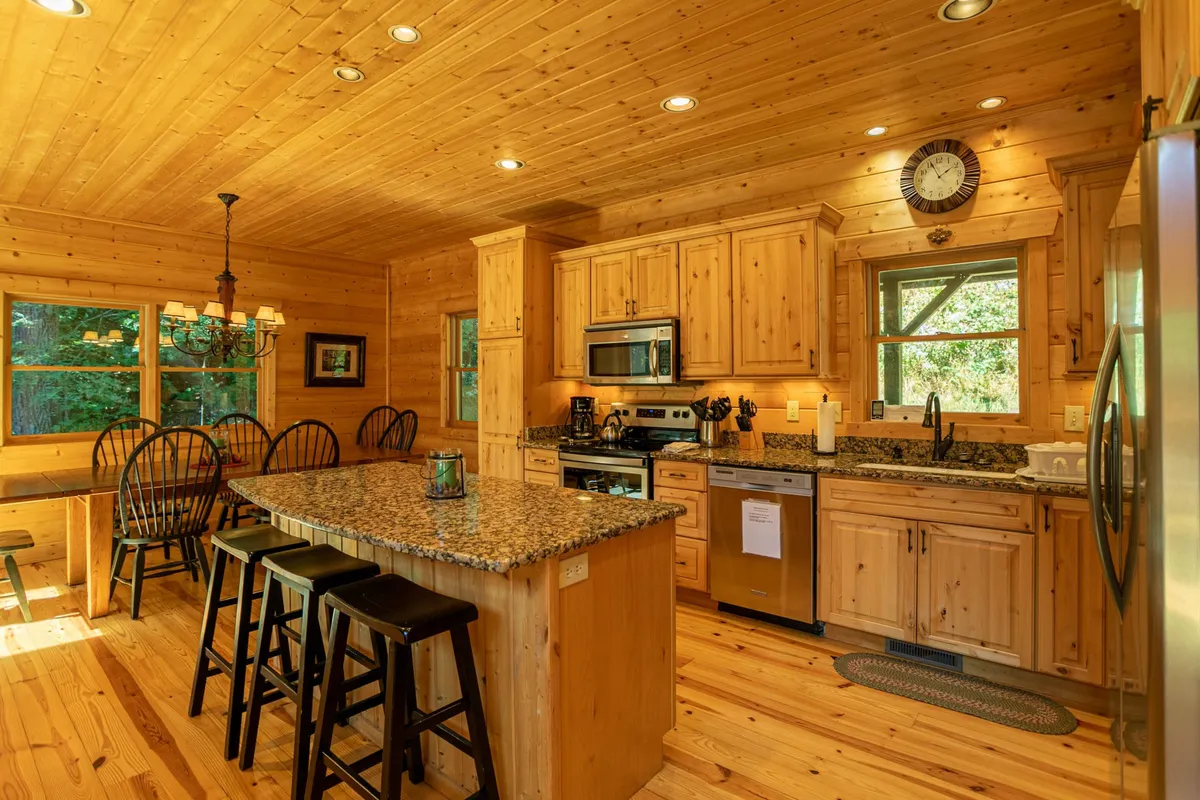 A well-appointed kitchen with granite countertops, wooden cabinetry, and stainless steel appliances, adjacent to a cozy dining area with black chairs. The natural light pouring in through the windows enhances the welcoming mood, and both the kitchen and dining area are visible.