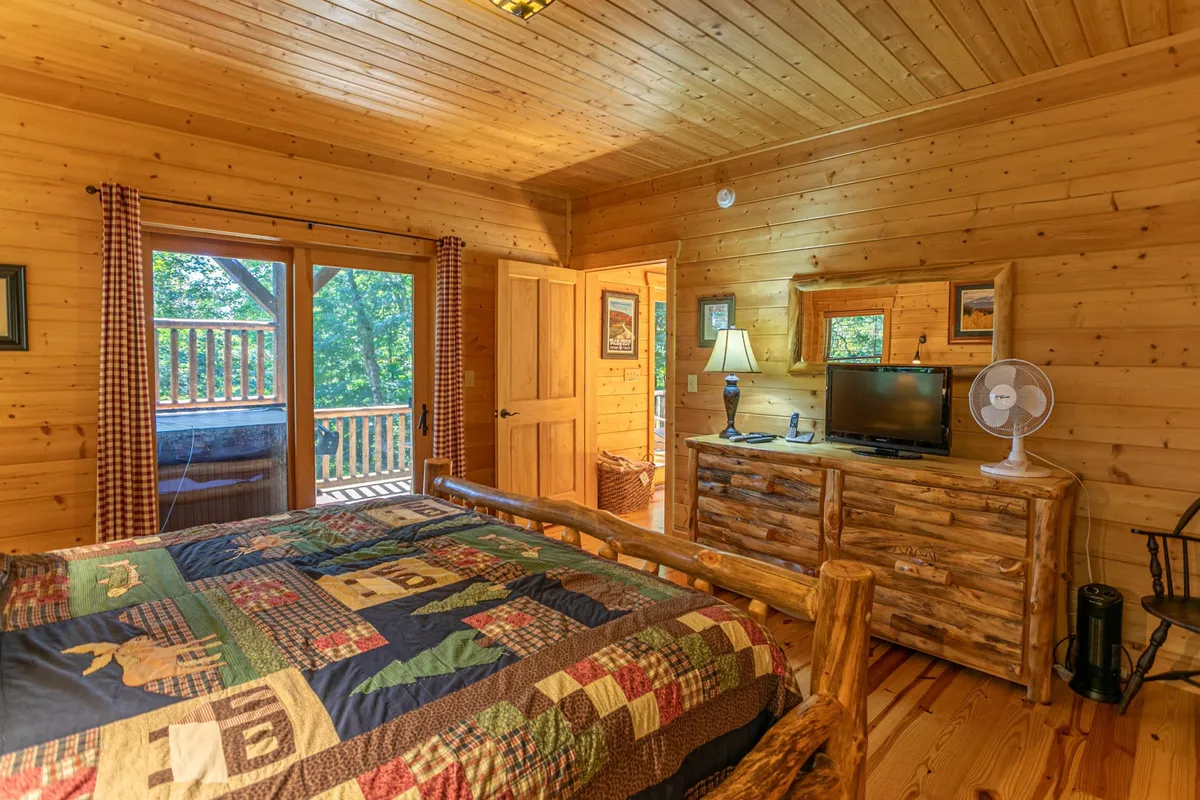This image displays a warmly lit bedroom featuring a bed with a quilt, wooden furniture, large windows, a TV, and a lamp. The room feels inviting and comfortable, with views to the outside and a relaxing atmosphere ideal for curling up and enjoying a movie marathon.