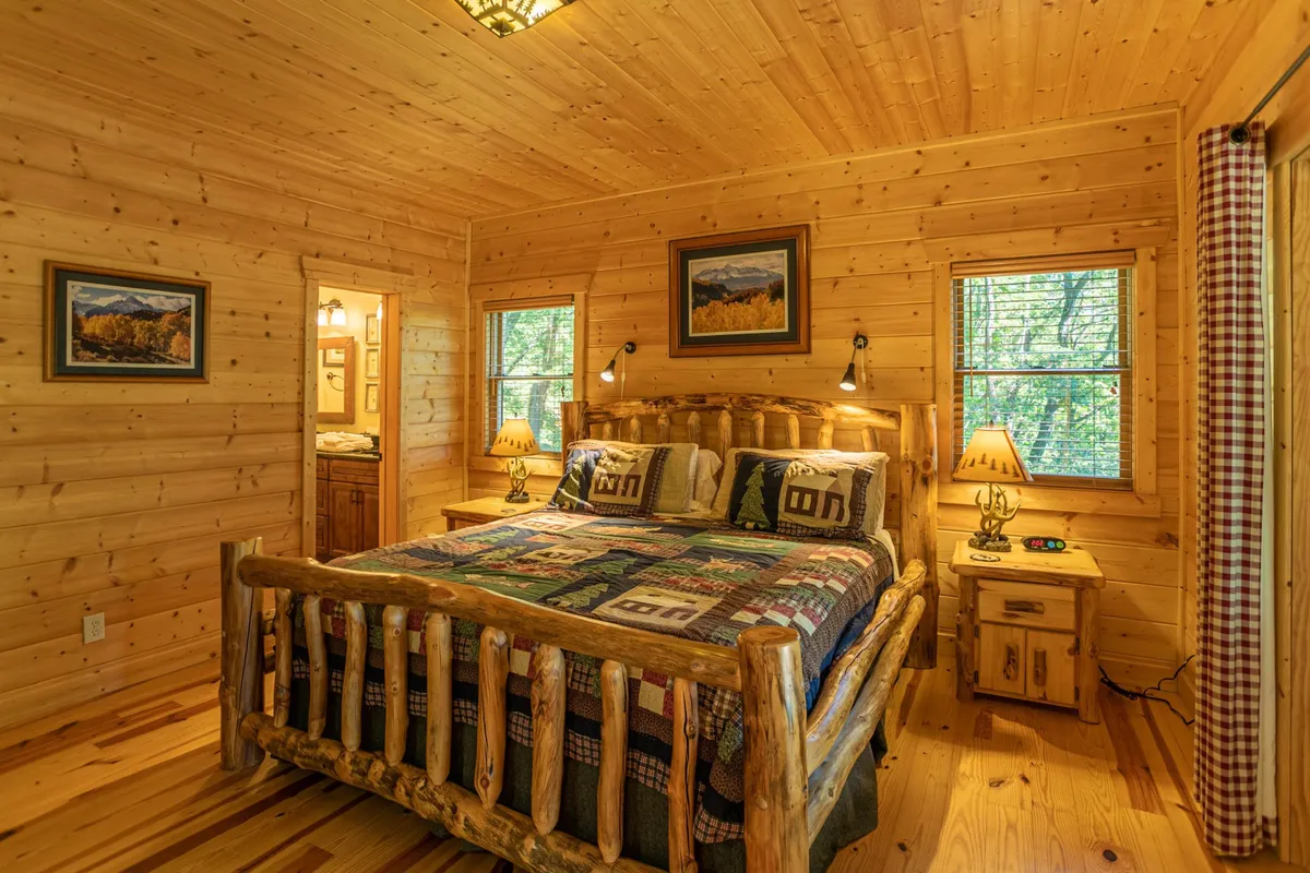 A warm and rustic bedroom featuring a log bed with a colorful quilt, wooden nightstands, framed artwork, and soft lighting from bedside lamps. The room exudes comfort and is designed for restful nights.