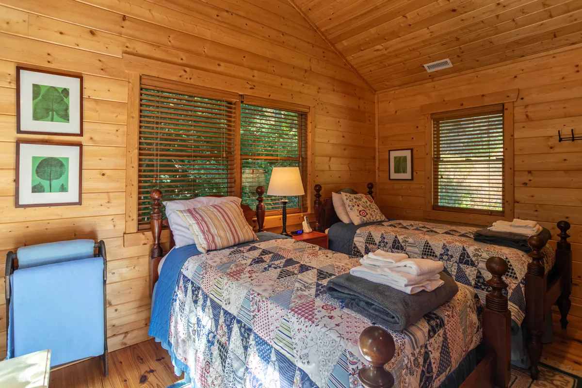 A tranquil alcove with two twin beds, accented by a table lamp, framed artwork, and cozy wooden walls. The arrangement hints at a comfortable spot for sleepovers or late-night chats, with homey touches and soft lighting.