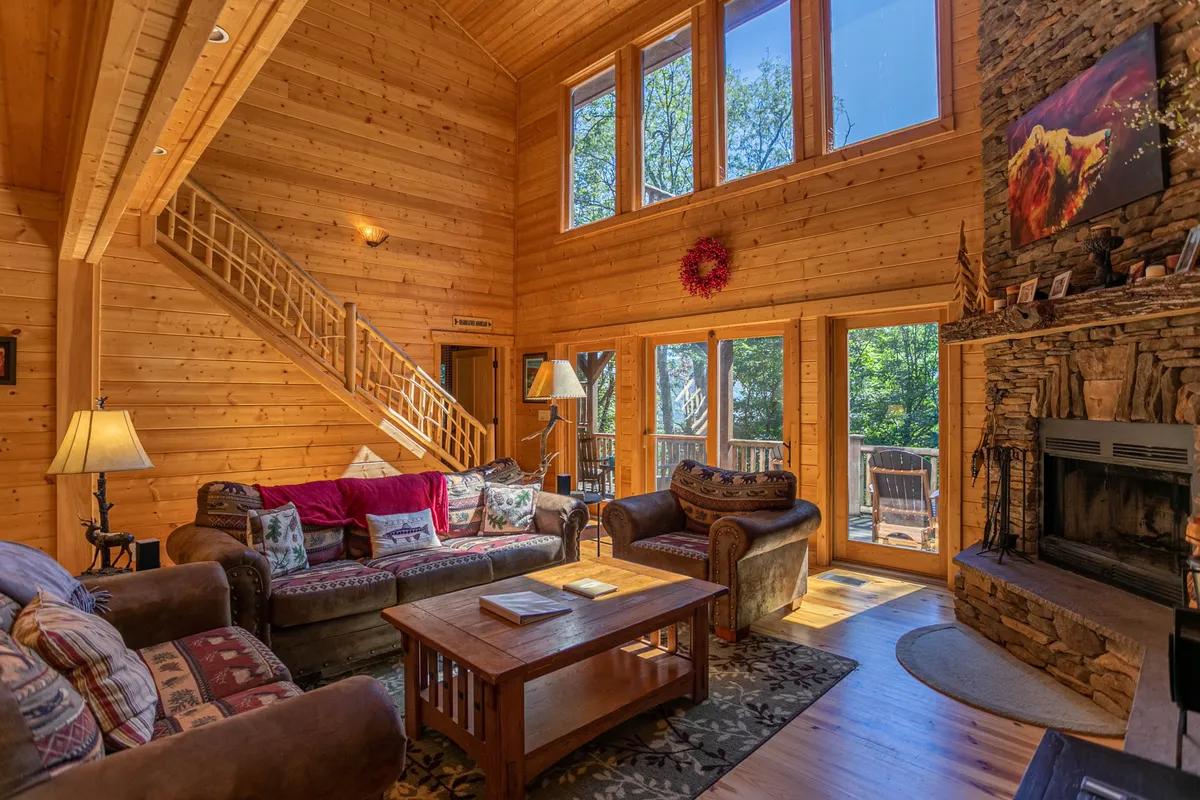A spacious living area featuring a dramatic stone fireplace rising two stories, comfortable sofas facing the fire, large windows framing breathtaking mountain views, a wooden staircase, and abundant natural light that fills the open-concept space.