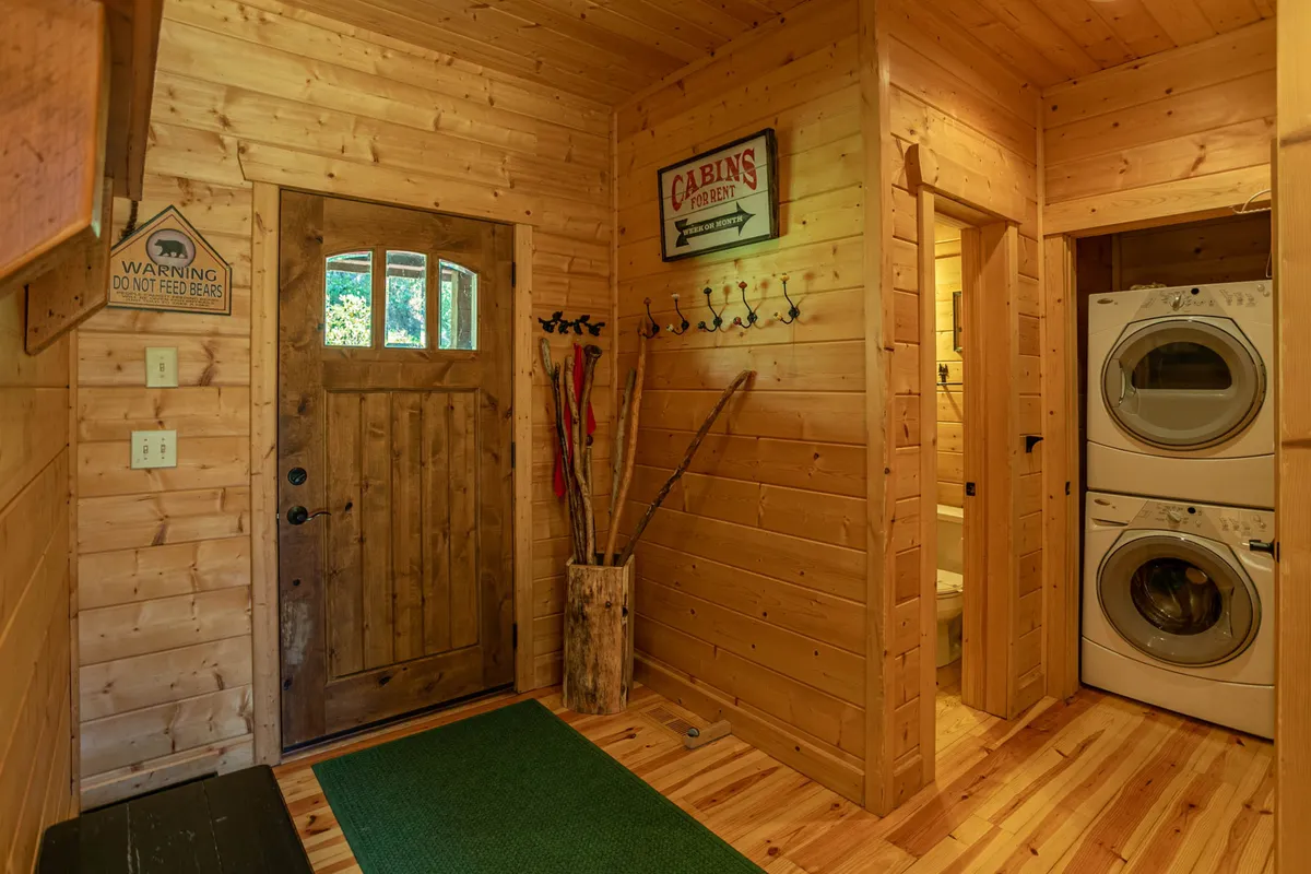 This image displays the entryway to the lodge, featuring a wooden door, laundry area, and hooks for outdoor gear. A warning sign is also visible, reinforcing the sense of adventure and preparedness required for mountain exploration.