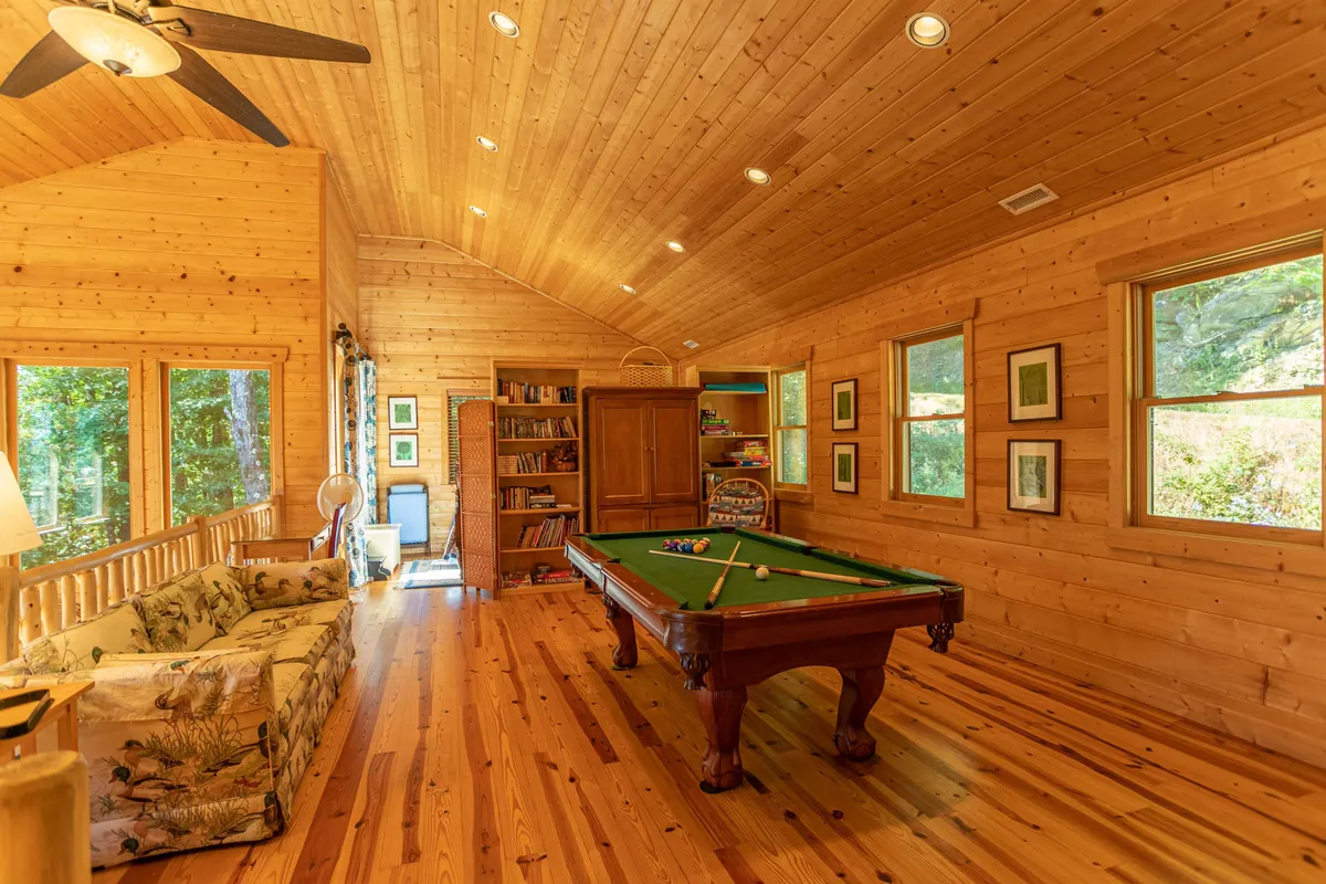 This image shows a cozy loft area featuring a pool table as the central element, surrounded by a comfortable sofa, a bookshelf, and windows letting in natural light. The arrangement suggests a multifunctional space for games, relaxation, and socializing.